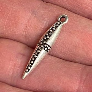 May include: A silver-colored pendant with a pointed shape. The pendant has a loop at the top for hanging and is decorated with small, raised dots and a cross-like pattern. The overall design is simple yet elegant.