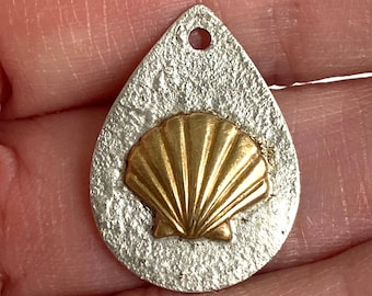 May include: A silver-toned pendant in a teardrop shape with a textured surface. A gold-colored seashell is centered on the pendant. The pendant has a small hole at the top for hanging. Jewelry.