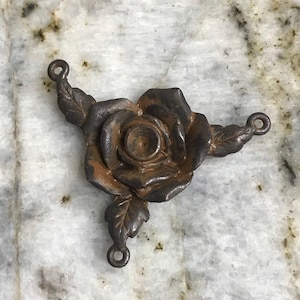 May include: A detailed, antique-style metal rose pendant with three leaves and three loops for attaching to a necklace or other craft project. The rose is a dark, weathered brown color.