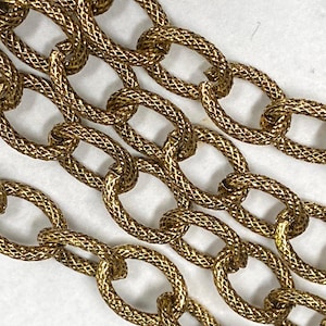 May include: Close-up of a gold-colored chain with an intricate woven pattern. The chain links are oval-shaped and interlocked, creating a textured surface. The chain is displayed against a white background, highlighting its metallic sheen. This jewelry piece is suitable for necklaces or bracelets.