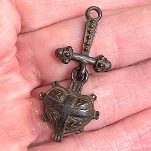 Puede incluir: Un pequeño colgante de estilo antiguo con una cruz y un corazón. El colgante está hecho de un metal oscuro con detalles intrincados. El corazón tiene un diseño de cruz y acentos de rayos. La cruz tiene extremos decorativos y un lazo para colgar.