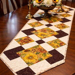 May include: A quilted table runner with a repeating pattern of yellow sunflowers and brown squares. The runner is on a dark wooden table, with a floral arrangement and candle holders in the background. The runner is approximately 14 inches wide.