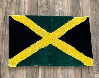 Pode incluir: Um tapete retangular com o design da bandeira jamaicana. O tapete tem um canto superior esquerdo e inferior direito preto, um fundo verde e uma cruz amarela no centro. Está colocado sobre uma superfície de madeira.