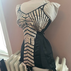 May include: A black corset with gold sequin embellishments in a geometric pattern. The corset has adjustable straps and garters.