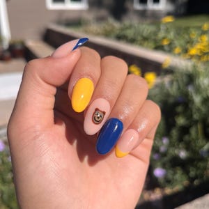 May include: A hand with oval-shaped nails, each painted in a different design. Colors include yellow, blue, and a nude base with a cartoon character. One nail has a blue diagonal stripe, and another has a yellow tip.
