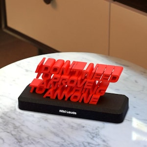 May include: Red 3D printed quote on a black base that reads "I don't need to prove anything to anyone" with the name "Niki Lauda" below the quote. The quote is on a white marble table.