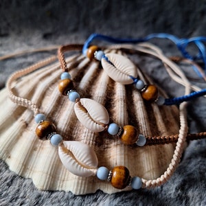 Könnte beinhalten: Drei übereinanderliegende Halsketten mit braunen Schnüren und weißen Muscheln. Jede Halskette hat blaue und braune Perlen.