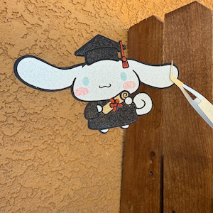 Puede incluir: Un gorro y una túnica de graduación blancos y negros con una cinta roja y un rollo. El personaje lleva un gorro de graduación negro con un borla roja. El personaje sostiene un rollo en su pata derecha. El personaje es un perro de dibujos animados blanco y negro con purpurina.