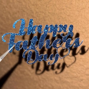 Puede incluir: Un adorno para pastel azul brillante con las palabras "Happy Father's Day" en una fuente cursiva. El adorno tiene forma de corazón en el diseño. La imagen está sobre un fondo marrón.