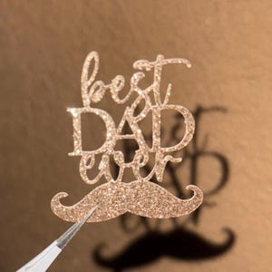 Puede incluir: Un adorno para pastel dorado brillante con las palabras "best DAD ever" y un diseño de bigote. El adorno para pastel está sujeto con pinzas sobre un fondo marrón. El texto está en una fuente cursiva.