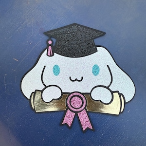 May include: A glittery light blue cartoon character wearing a black graduation cap with a pink tassel. The character is holding a gold scroll with a pink ribbon. The eyes are turquoise.