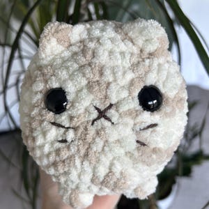 May include: A crocheted plush cat toy with a white and beige striped pattern. The cat has black eyes and a stitched brown nose.