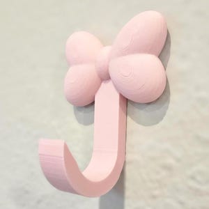 May include: A light pink wall hook shaped like a bow. The bow is at the top, with a curved hook extending downwards. The hook is mounted on a light-colored wall. The bow and hook are made of a solid material.