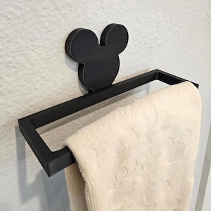 May include: Black metal towel rack featuring a Mickey Mouse head design. The rack is rectangular and holds a cream-colored towel. The rack is mounted on a white wall, adding a touch of whimsy to bathroom decor.