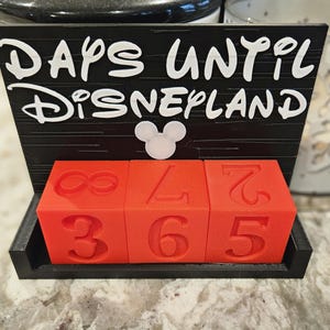 May include: A black countdown calendar with the white text DAYS UNTIL DISNEYLAND and a white mouse head icon. The base holds three red cubes with embossed numbers, including an infinity symbol, a 3, a 6, a 5, a 7, and a 2, used to track time.