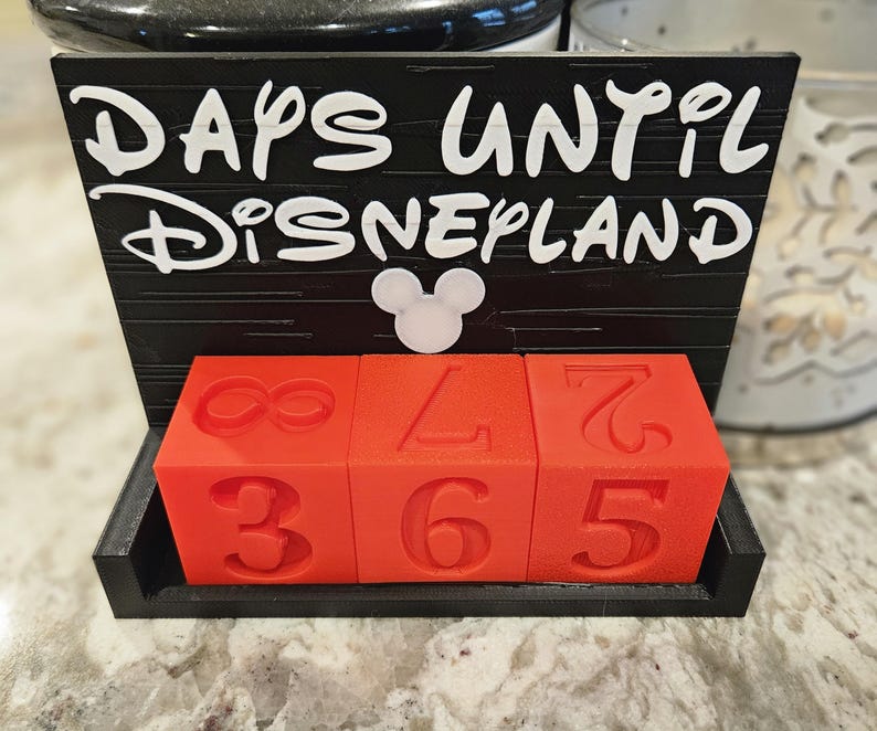 May include: A black countdown calendar with the white text DAYS UNTIL DISNEYLAND and a white mouse head icon. The base holds three red cubes with embossed numbers, including an infinity symbol, a 3, a 6, a 5, a 7, and a 2, used to track time.