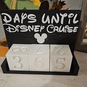May include: A black rectangular sign with white text that reads DAYS UNTIL DISNEY CRUISE and a white Mickey Mouse icon. Below the sign are three white cubes with embossed numbers, arranged in a black base to display a countdown of days.