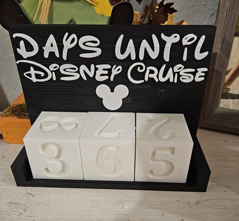 May include: A black rectangular sign with white text that reads DAYS UNTIL DISNEY CRUISE and a white Mickey Mouse icon. Below the sign are three white cubes with embossed numbers, arranged in a black base to display a countdown of days.