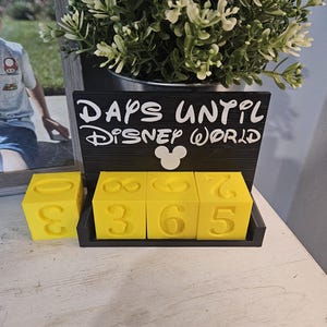 May include: A black countdown calendar with the white text "DAYS UNTIL DISNEY WORLD" and a white Mickey Mouse icon. The calendar features three bright yellow cubes with embossed numbers, allowing for the display of a three-digit number count.