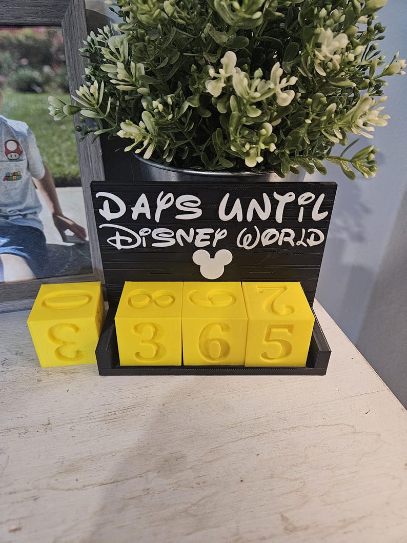 May include: A black countdown calendar with the white text "DAYS UNTIL DISNEY WORLD" and a white Mickey Mouse icon. The calendar features three bright yellow cubes with embossed numbers, allowing for the display of a three-digit number count.