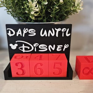 May include: A black countdown sign with white text that reads DAYS UNTIL DISNEY, featuring a Mickey Mouse icon. Three red cubes with embossed numbers sit in the base, displaying 365, with an additional red cube showing the number 1.