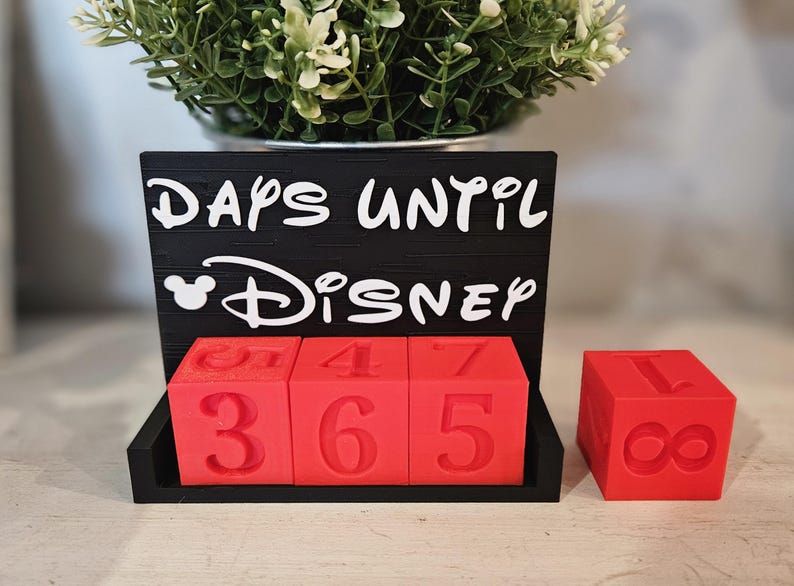 May include: A black countdown sign with white text that reads DAYS UNTIL DISNEY, featuring a Mickey Mouse icon. Three red cubes with embossed numbers sit in the base, displaying 365, with an additional red cube showing the number 1.