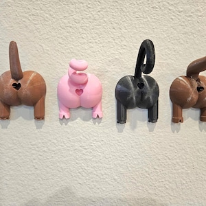 May include: Four novelty butt-shaped wall decorations in brown, pink, black, and brown. Each has a heart-shaped cutout and a tail. The objects are mounted on a textured, off-white wall.