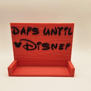 May include: A rectangular red countdown sign with a small shelf at the base. The sign features black embossed text that reads DAYS UNTIL DISNEY, with a small Mickey Mouse head icon integrated into the text. The surface has a horizontal line texture.