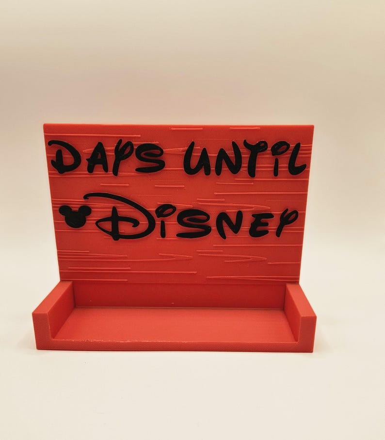 May include: A rectangular red countdown sign with a small shelf at the base. The sign features black embossed text that reads DAYS UNTIL DISNEY, with a small Mickey Mouse head icon integrated into the text. The surface has a horizontal line texture.