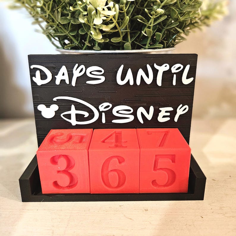May include: A black countdown calendar with white text that reads DAYS UNTIL DISNEY, featuring a mouse ear icon. The calendar includes three red square blocks with embossed numbers, currently displaying the digits 3, 6, and 5 on the front faces.