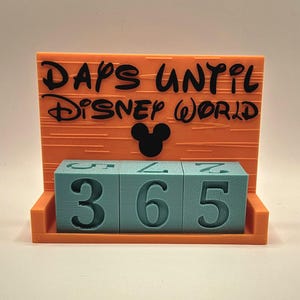 May include: An orange countdown calendar with a black silhouette of a mouse head and the text "DAYS UNTIL DISNEY WORLD" printed in black. Three light blue square blocks with dark blue numbers are arranged in a row to display the number 365.