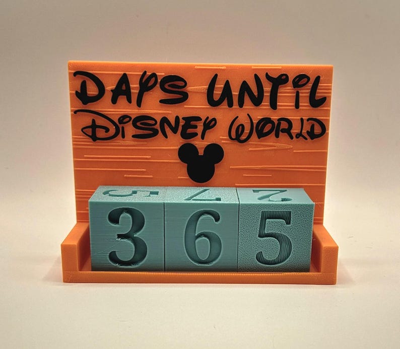 May include: An orange countdown calendar with a black silhouette of a mouse head and the text "DAYS UNTIL DISNEY WORLD" printed in black. Three light blue square blocks with dark blue numbers are arranged in a row to display the number 365.