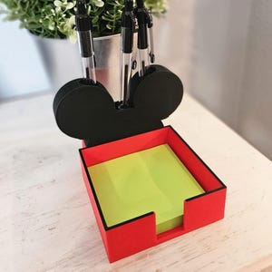 May include: A red and black desk organizer shaped like a mouse head. The organizer has a pen holder and a square compartment filled with yellow sticky notes. Several black pens are in the pen holder.