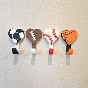 May include: Four heart-shaped wall hooks with sports ball designs. The hooks feature a soccer ball, football, baseball, and basketball. Each hook has a different color scheme, including black, white, brown, and orange. The hooks are mounted on a light gray wall.