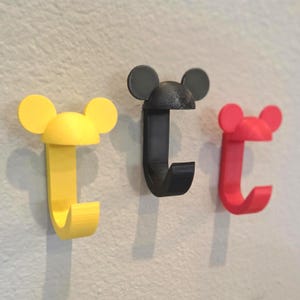 May include: Three wall hooks in yellow, black, and red, each shaped like a mouse head with rounded ears. The hooks are mounted on a light-colored wall, offering a playful and decorative storage solution.