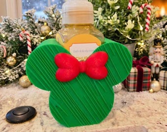 May include: A clear plastic soap dispenser bottle encased in a green, mouse-ear-shaped holder featuring a bright red bow. The label on the bottle reads Bath & Body Works. The holder has a textured, ridged surface and a white pump top.
