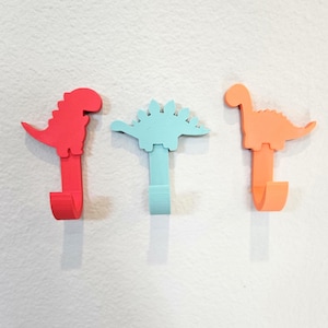 May include: Three colorful dinosaur-shaped wall hooks. The hooks are in red, light blue, and orange. Each hook has a dinosaur silhouette at the top and a curved hook at the bottom. The hooks are mounted on a white wall.