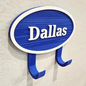 May include: A blue and white wall hook with the name "Dallas" in white lettering. The hook has a blue oval shape with a white border and two blue hooks below.