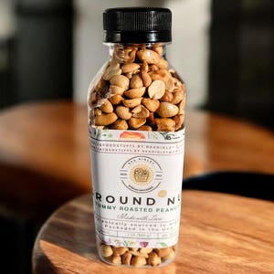 Puede incluir: Un frasco de vidrio transparente lleno de cacahuetes tostados. El frasco tiene una tapa negra y una etiqueta blanca que dice "Round Nut Yummy Roasted Peanuts Made with Love" y "Ethically sourced in Nigeria Packaged in the USA 1.2 lb (400 g)".