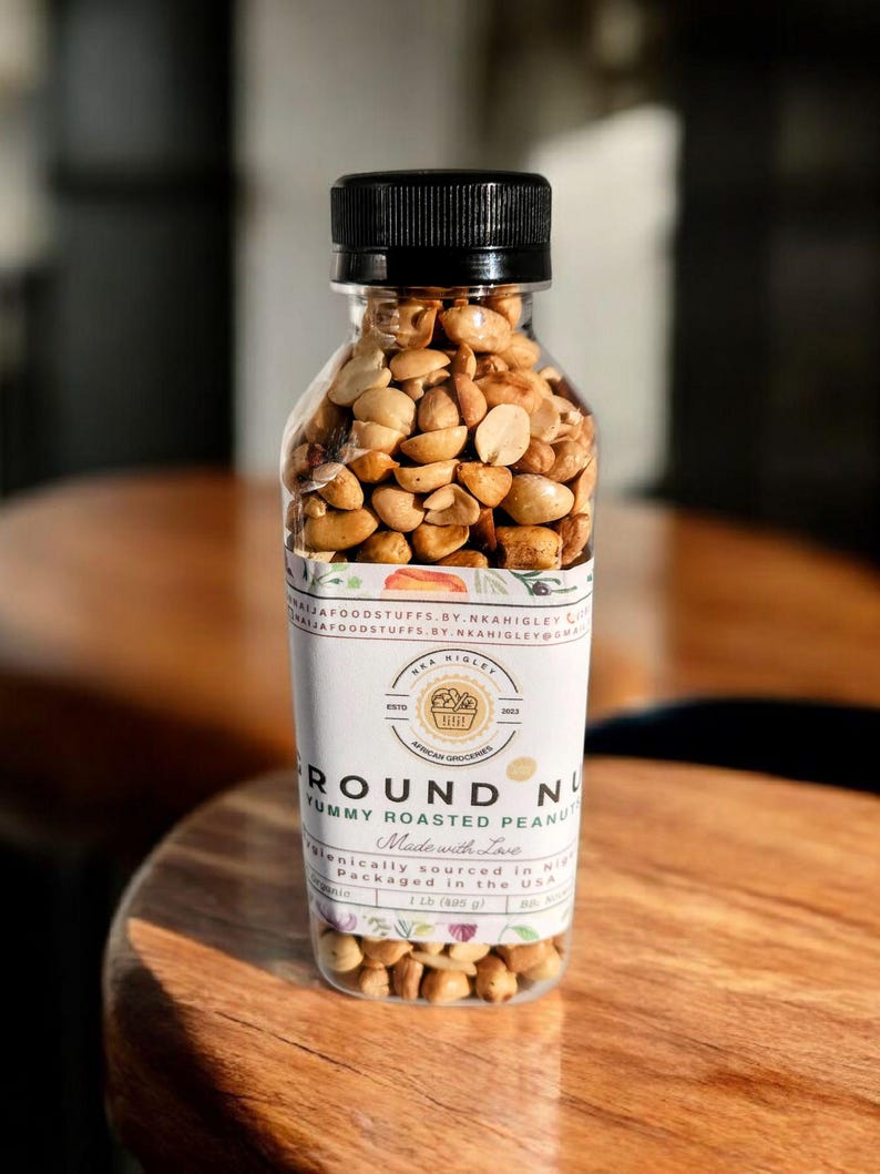 Puede incluir: Un frasco de vidrio transparente lleno de cacahuetes tostados. El frasco tiene una tapa negra y una etiqueta blanca que dice "Round Nut Yummy Roasted Peanuts Made with Love" y "Ethically sourced in Nigeria Packaged in the USA 1.2 lb (400 g)".