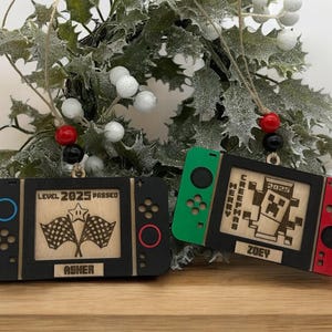 May include: Two wooden Christmas ornaments shaped like game consoles. One is black with blue and red accents, the other is green and red. Both have engraved details and the names "Asher" and "Zoey". The ornaments are hanging in front of a festive background.