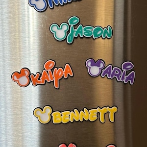Six colorful magnetic custom nameplates with Mouse ears. They are displayed are on a silver refrigerator. The girl names will come with a bow on the mouse ears. The boy names will not have a bow.