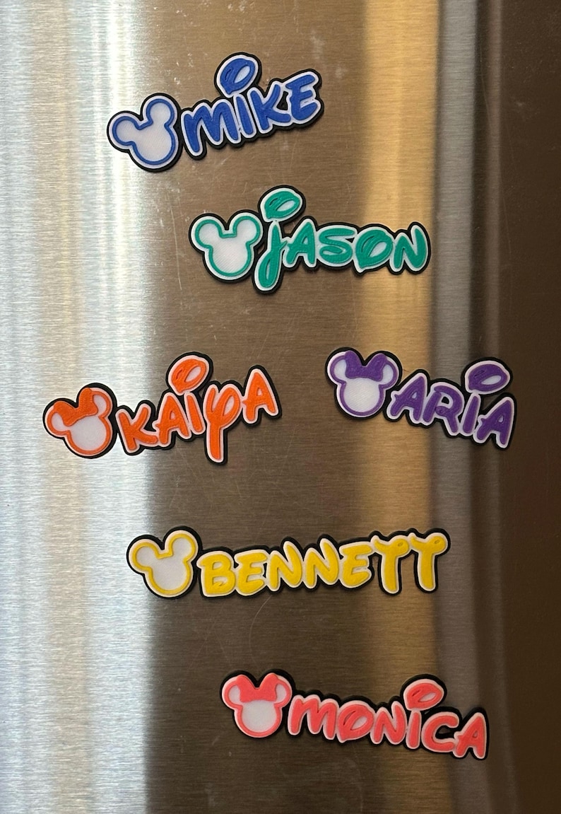 Six colorful magnetic custom nameplates with Mouse ears. They are displayed are on a silver refrigerator. The girl names will come with a bow on the mouse ears. The boy names will not have a bow.