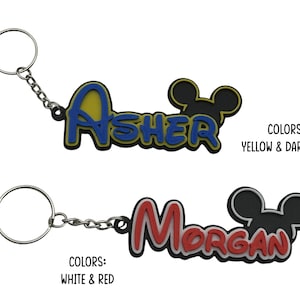 Two personalized keychains with a Mouse ear design. The first keychain is yellow and dark blue with the name 'ASHER' and the second keychain is white and red with the name 'Morgan'. The mouse head design on these do not have bows as they are the boy version keychain.