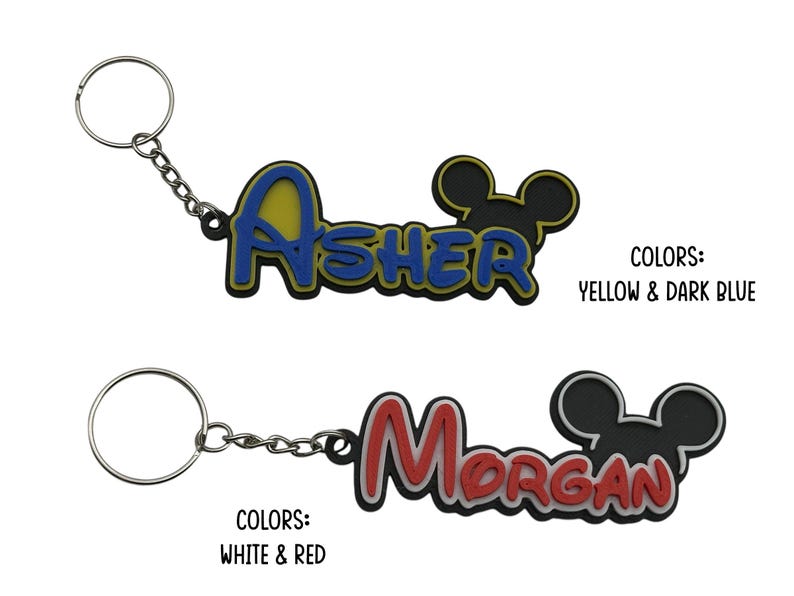 Two personalized keychains with a Mouse ear design. The first keychain is yellow and dark blue with the name 'ASHER' and the second keychain is white and red with the name 'Morgan'. The mouse head design on these do not have bows as they are the boy version keychain.