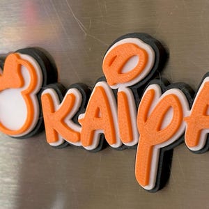 A 3D printed name tag with the name 'Kaiya' in orange letters with white outlines. The name is surrounded by a overall black outline. The tag also includes a small orange Mouse head with a bow. With three layers, this magnet is sturdy yet sleek looking.