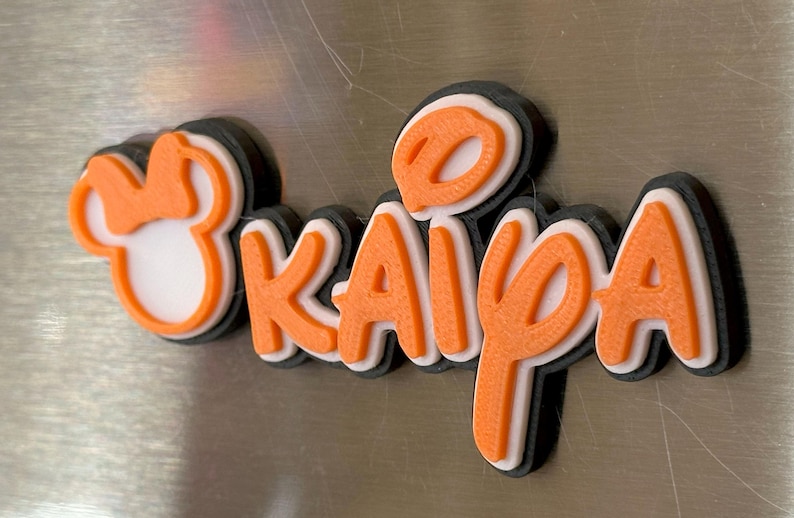 A 3D printed name tag with the name 'Kaiya' in orange letters with white outlines. The name is surrounded by a overall black outline. The tag also includes a small orange Mouse head with a bow. With three layers, this magnet is sturdy yet sleek looking.