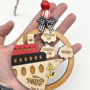 A hand holding one wooden circle ornament with a cruise boat image and 3 port locations clouds in this version. The ornament comes with 2 wooden beads, a black and white mini fabric bow and twine for hanging.