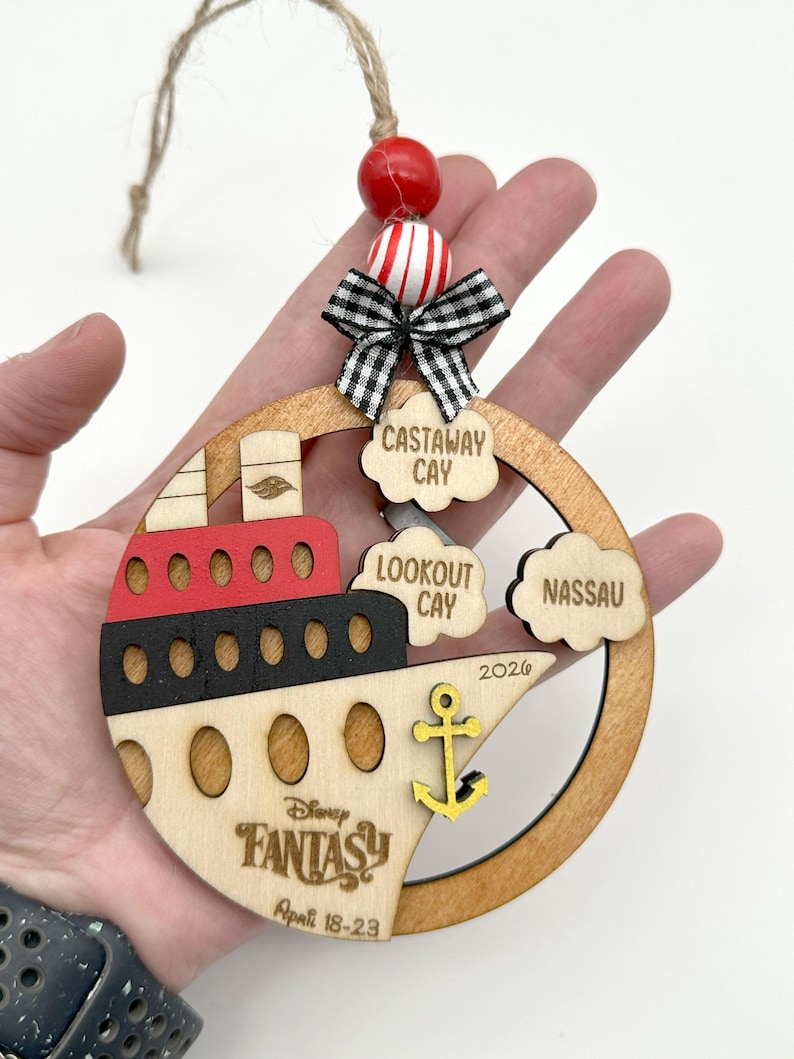 A hand holding one wooden circle ornament with a cruise boat image and 3 port locations clouds in this version. The ornament comes with 2 wooden beads, a black and white mini fabric bow and twine for hanging.