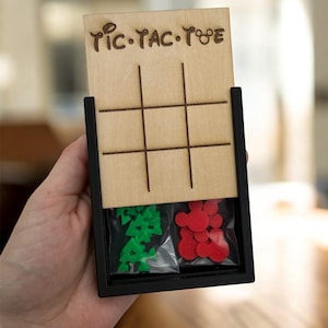 May include: Wooden Tic-Tac-Toe game with a black frame. The game board is light brown with etched lines and the words "Tic-Tac-Toe". The game pieces are green trees and red Mickey Mouse shapes, stored in clear bags.